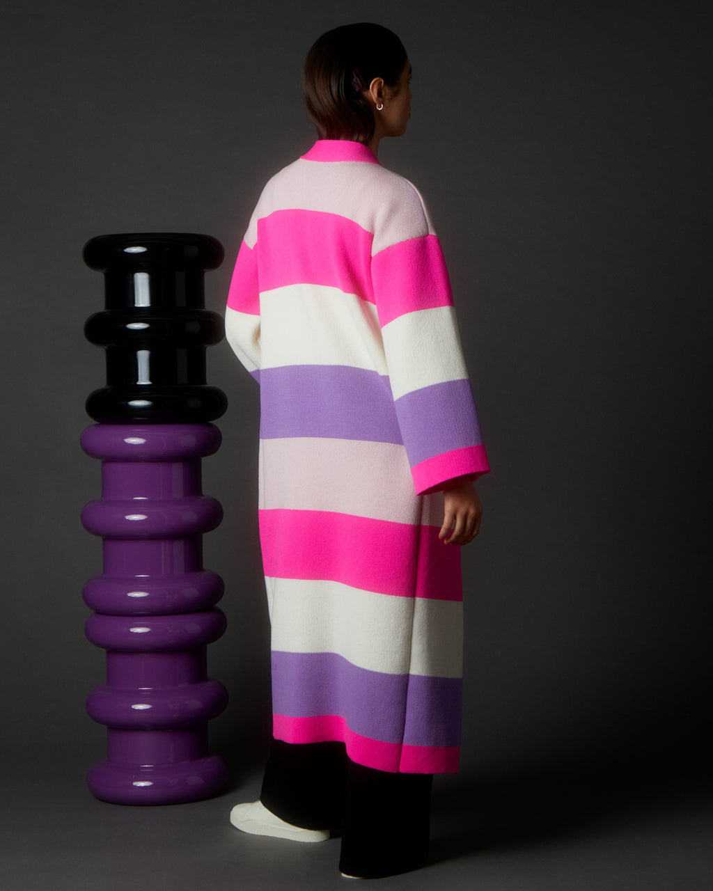 Person wearing a Luxurious Multicolour Striped Long Cardigan made with Wool & Cashmere Blend | Sustainable & Cozy standing next to a black and purple cylindrical sculpture on a dark background