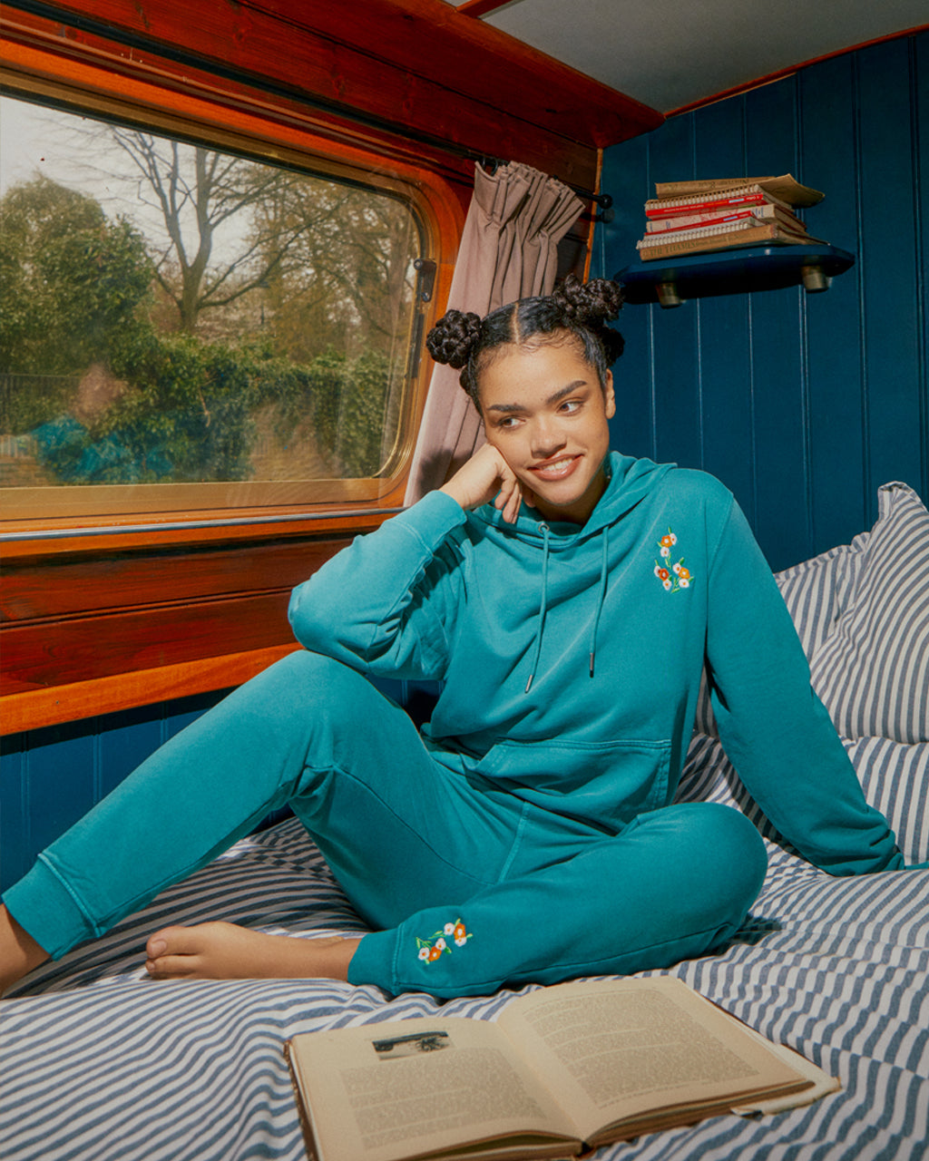 Person wearing a teal hoodie and Luxurious Floral Embroidered Joggers in Teal - 100% Organic Cotton sitting on a bed with an open book, in a room on a barge with blue walls and a window.