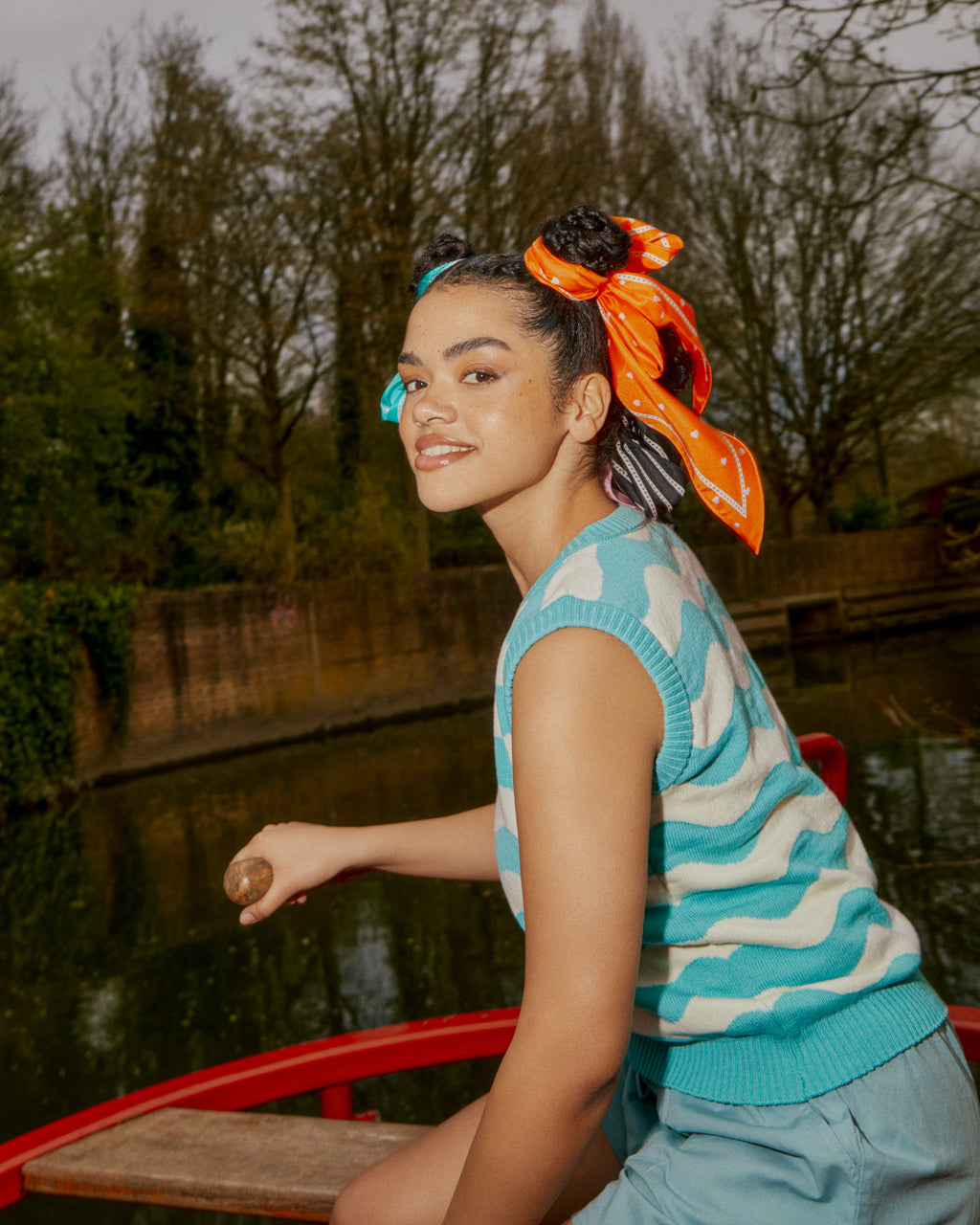 Woman with her hair tied up with a Tiny Hearts Long Orange Silk Neck Scarf sitting on a red barge by a body of water with trees in the background