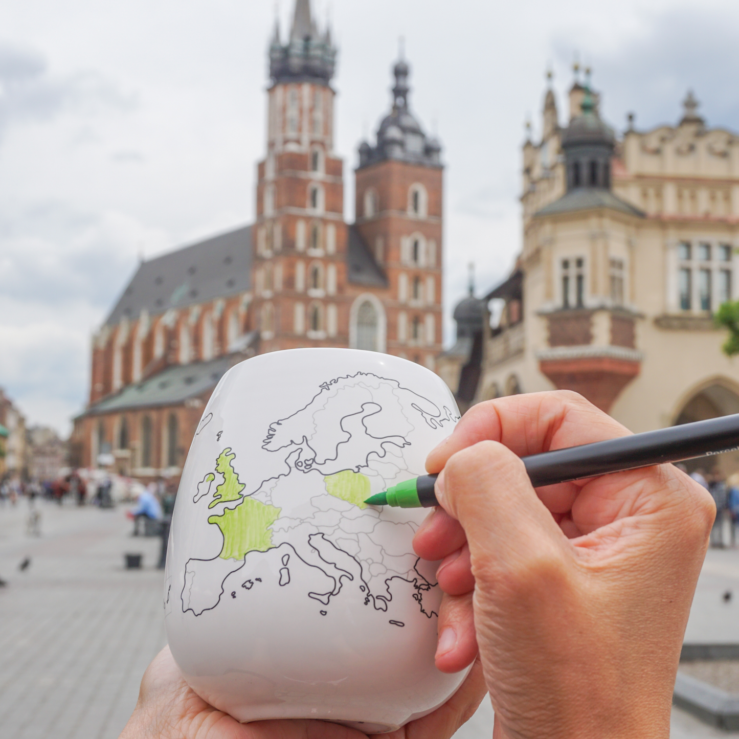 Person outside drawing in a european map on a ceramic mug with a cityscape in the background 