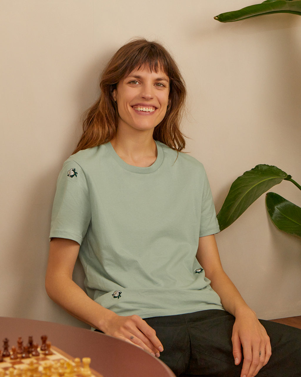 Woman wearing a Organic Cotton Buds Embroidered T-Shirt - Soft, Sustainable Fashion in Aloe Green | Eco-Friendly Apparel, sitting against a beige wall with a plant.