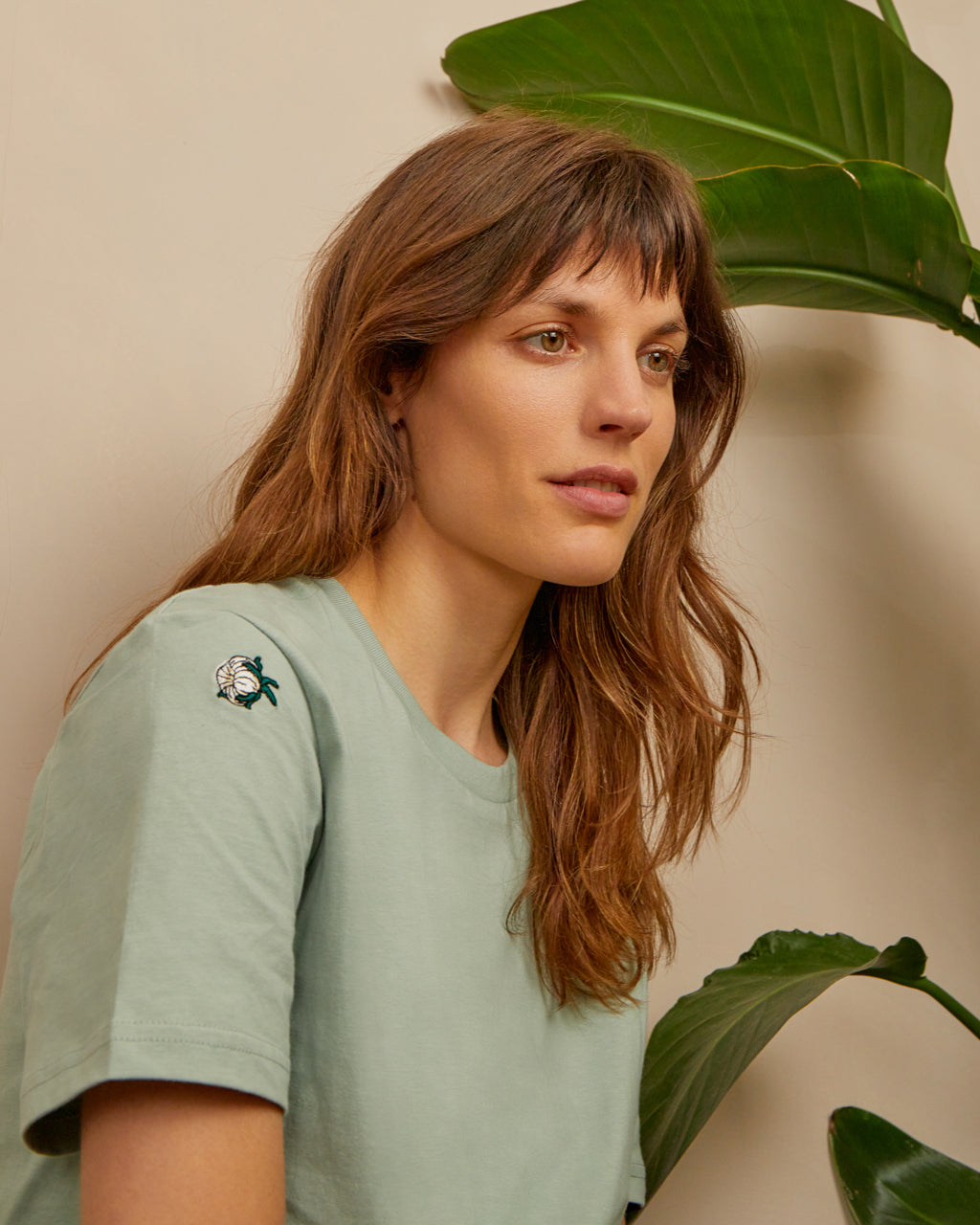 Woman wearing a Organic Cotton Buds Embroidered T-Shirt - Soft, Sustainable Fashion in Aloe Green | Eco-Friendly Apparel, standing against a beige wall with plants.