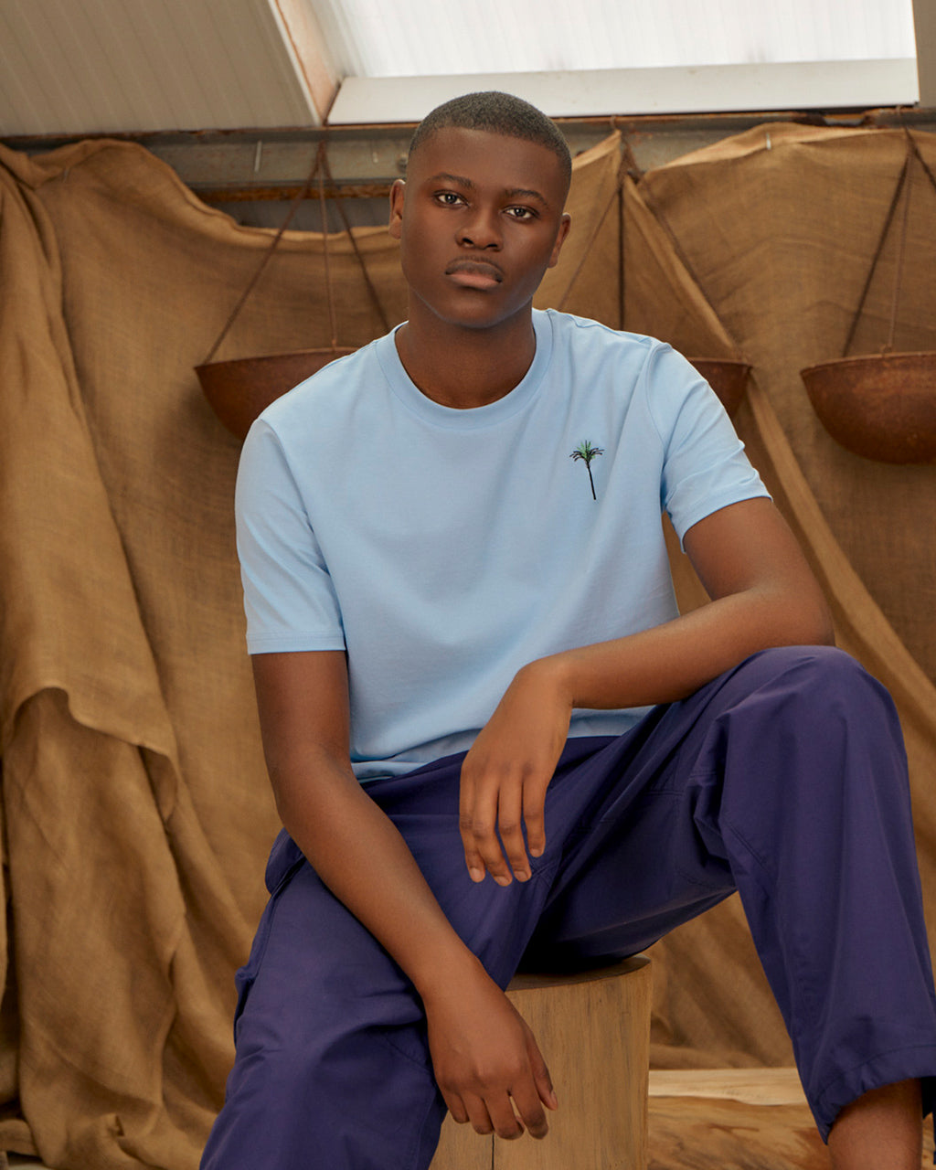 Person wearing a Palm Embroidered Organic Cotton Soft Blue T-Shirt and purple pants sitting against a brown fabric backdrop.
