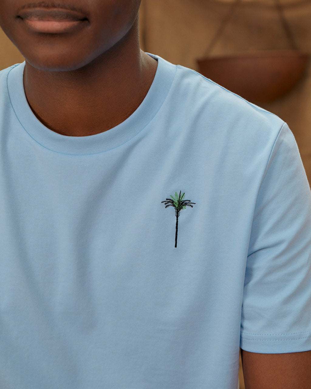 Person wearing a Palm Embroidered Organic Cotton Soft Blue T-Shirt on a blurred background.
