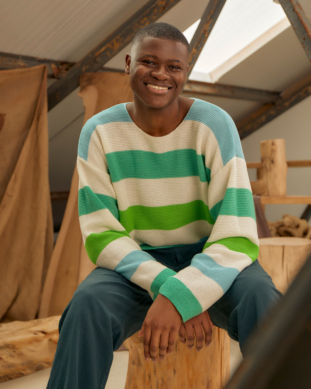 Man wearing a Eco-Friendly Striped Dropped Shoulder Top - Green & Blue | Wool & Cashmere Blend, Ethically Sourced sitting on a wooden stool indoors.