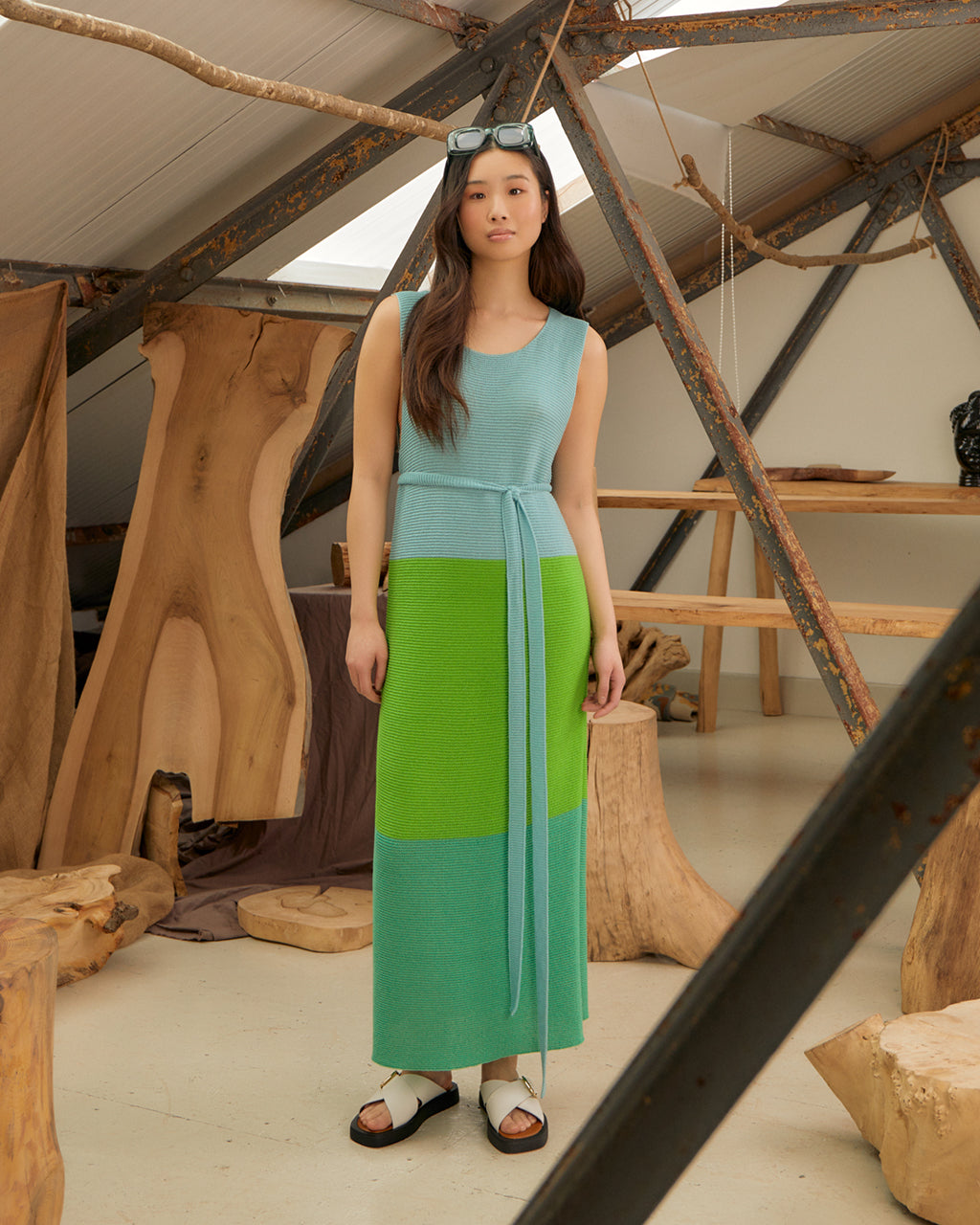 Woman wearing a Elegant Colour Block Belted Slit Dress in Green & Blue - Sustainable Wool & Cashmere, Made in London standing in a rustic indoor setting with wooden elements.