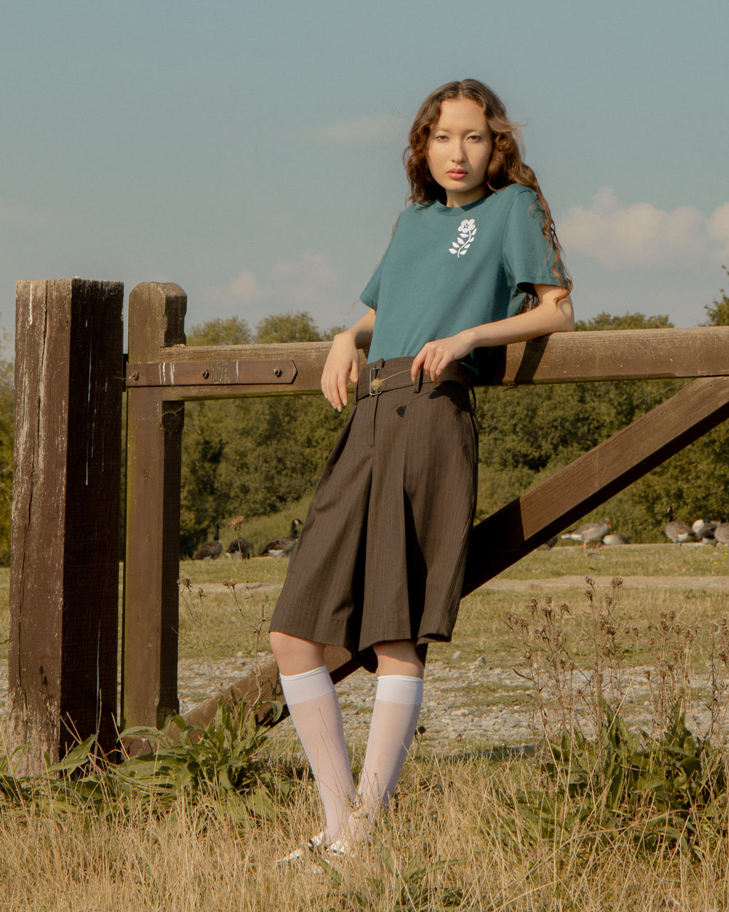 Woman wearing a Eco-Friendly Floral Embroidered Organic Cotton Teal T-Shirt and brown shorts standing by a wooden fence in a field with trees in the background