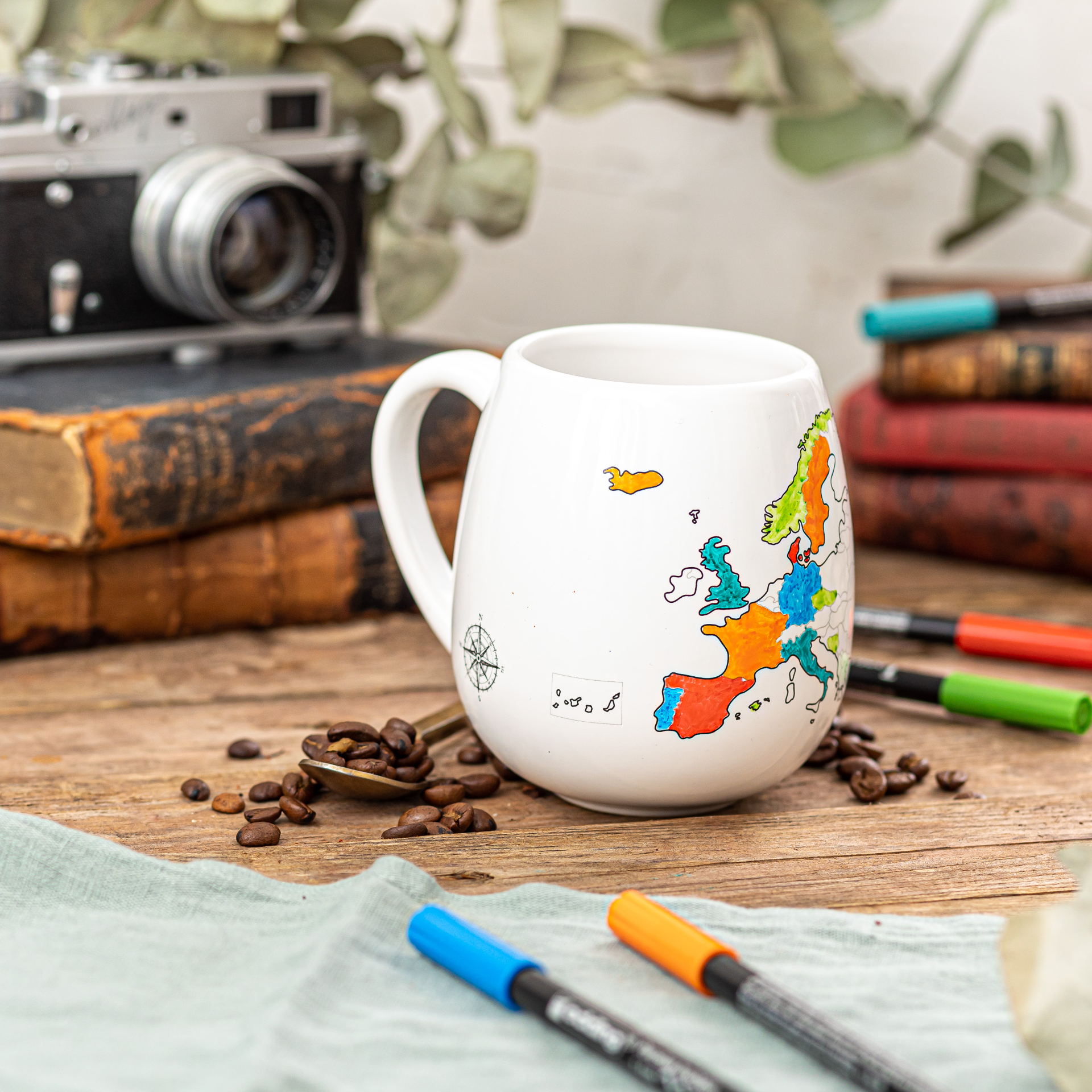 Ceramic white mug with a colorful map design on a wooden table with coffee beans and pens.