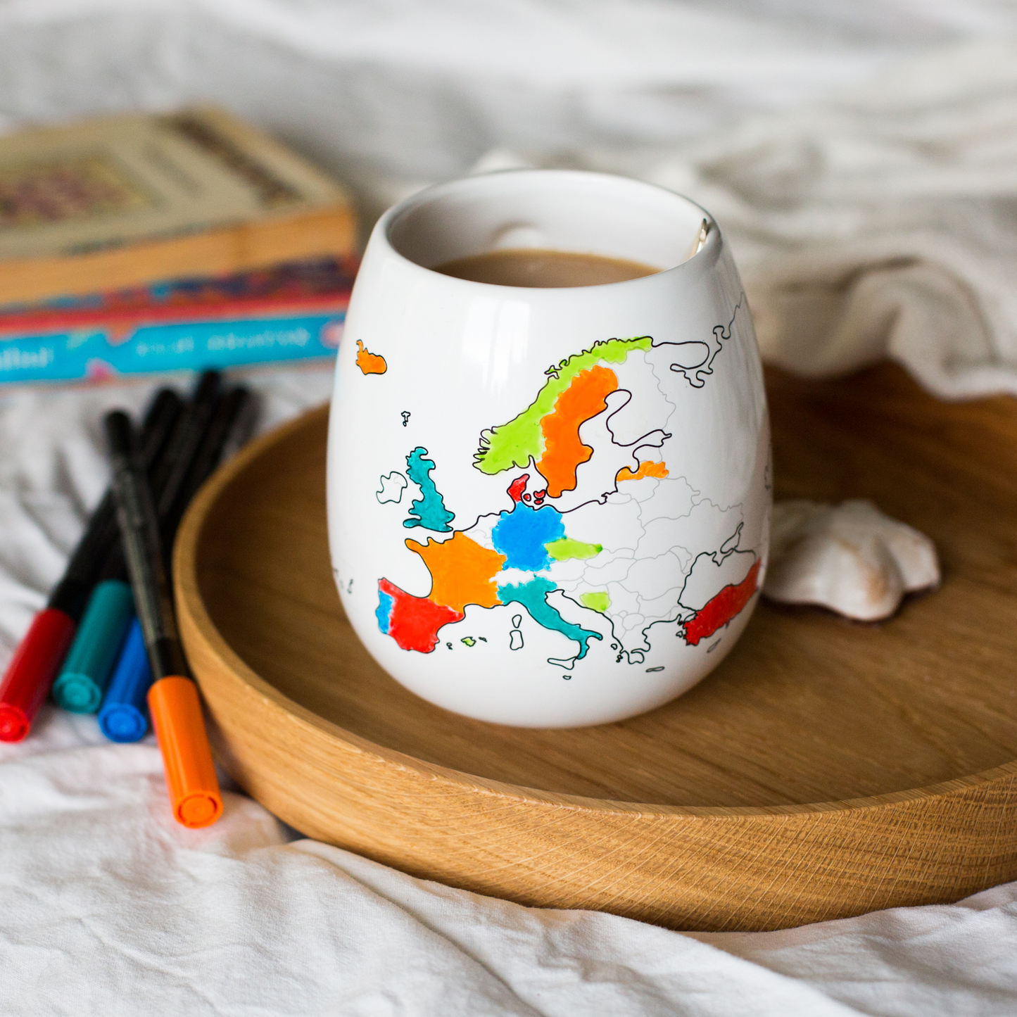 White mug with a colorful map of Europe design on a wooden tray with markers and books in the background.