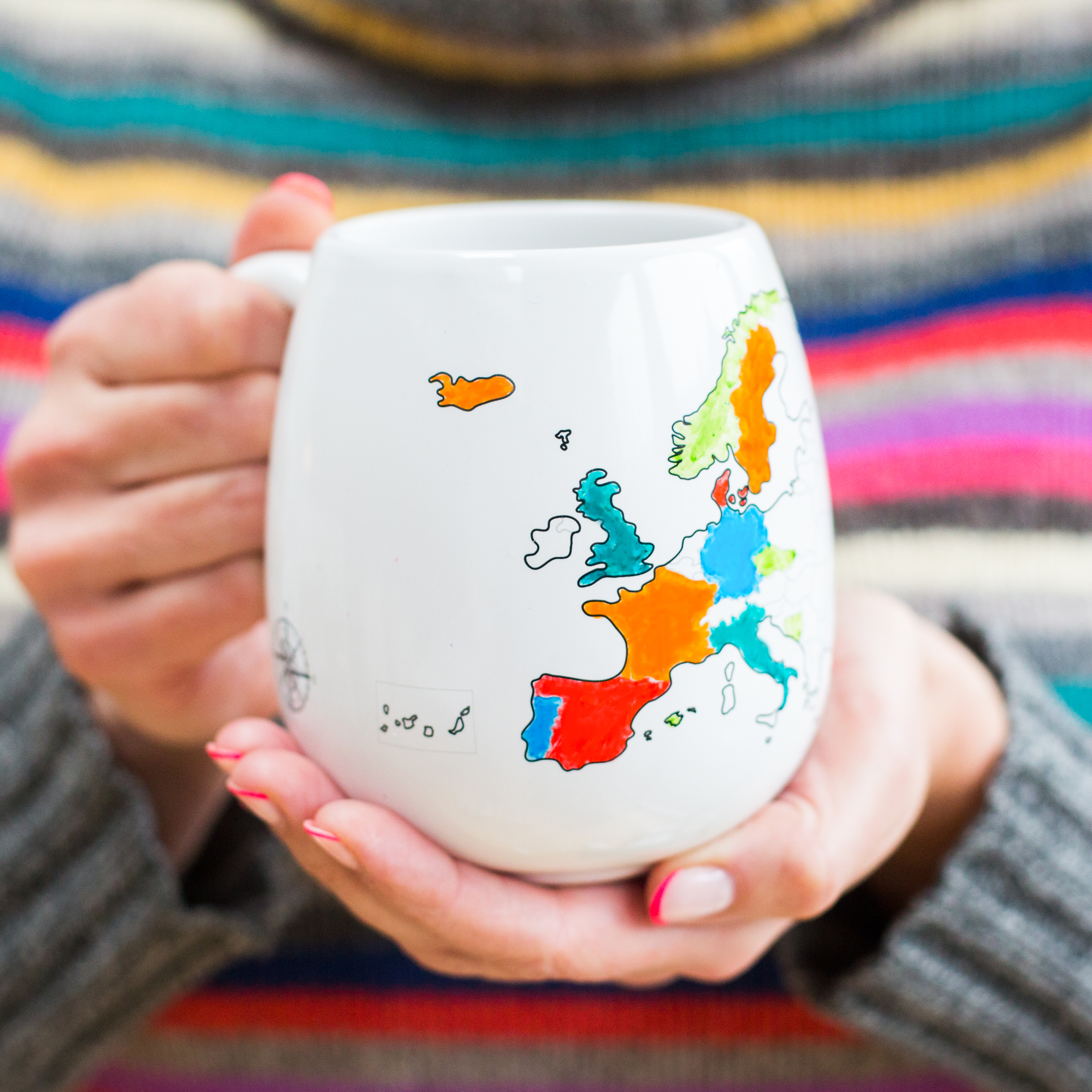 Person holding a mug with a colorful european map design earing a striped jumper in the background