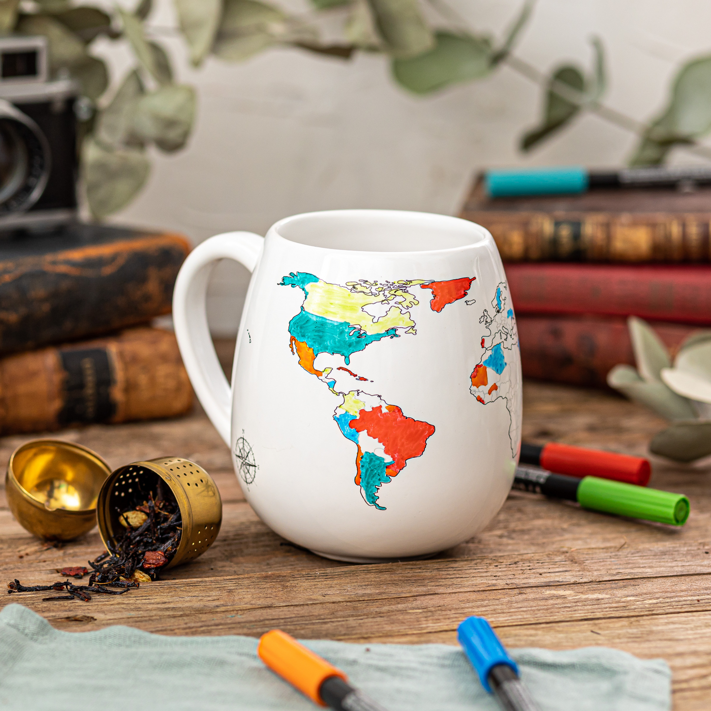 White mug with world map design coloured in on a wooden surface with books, stationery items and loose tea strainer