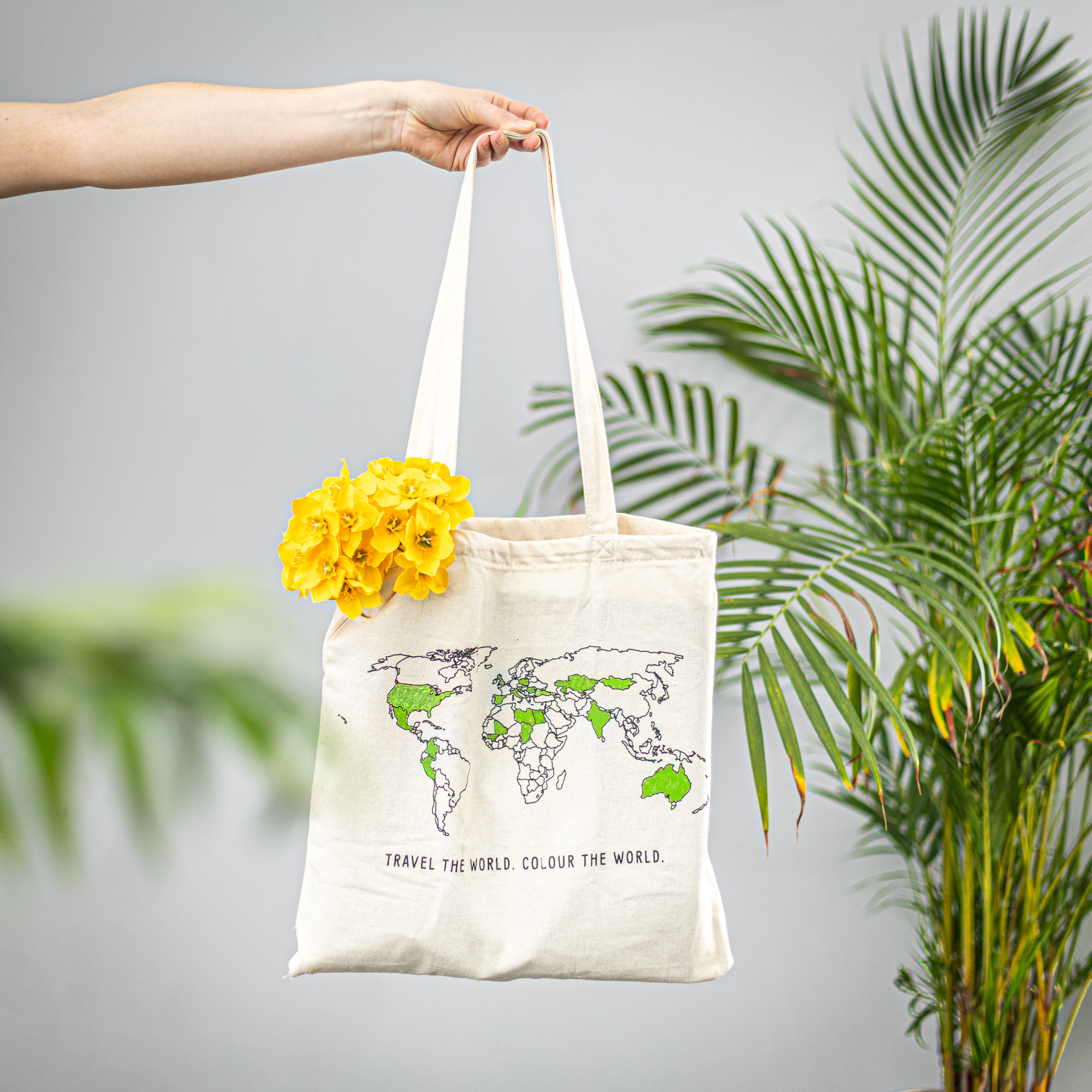 Tote bag with world map design held by a hand with yellow flowers, against a light gray background with green plants.
