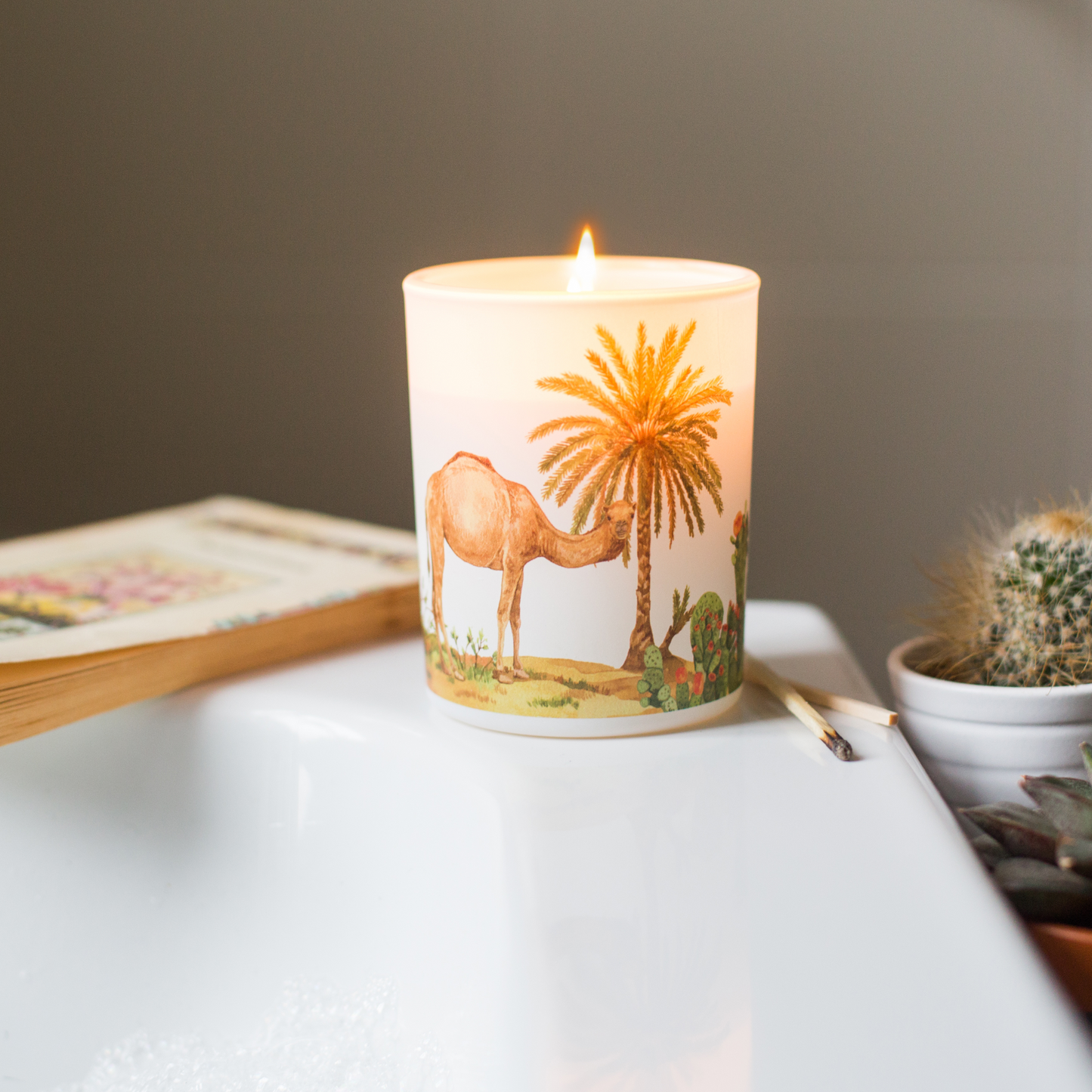 Candle with a desert scene design on a table next to a book and cactus plant.