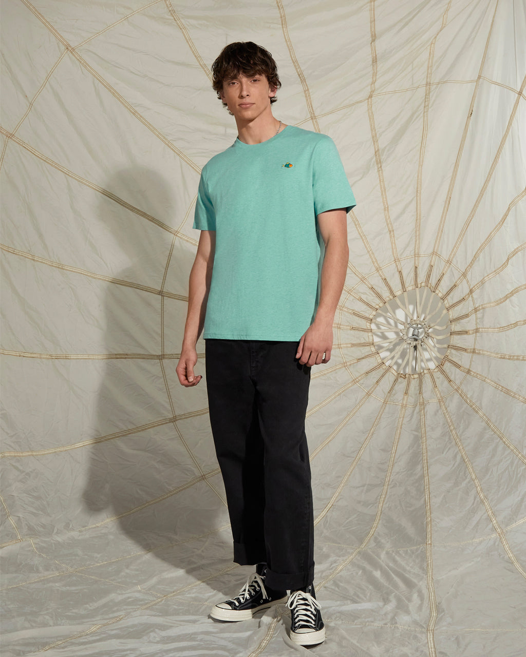 Man wearing a Heather Green - Super Soft Organic Cotton t-shirt and black pants standing against a decorative wall.