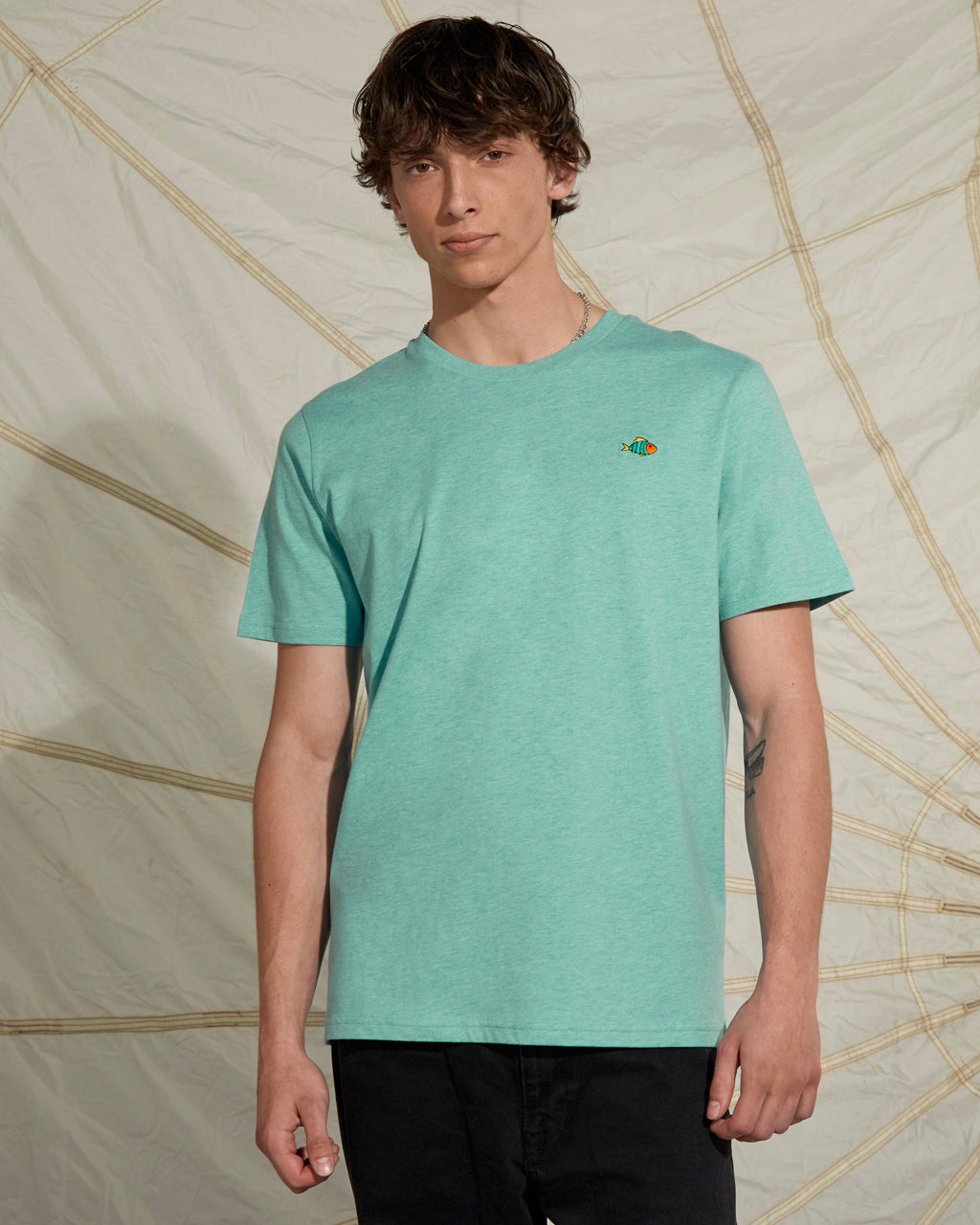 Man wearing a Heather Green - Super Soft Organic Cotton t-shirt with a small logo against a textured beige background