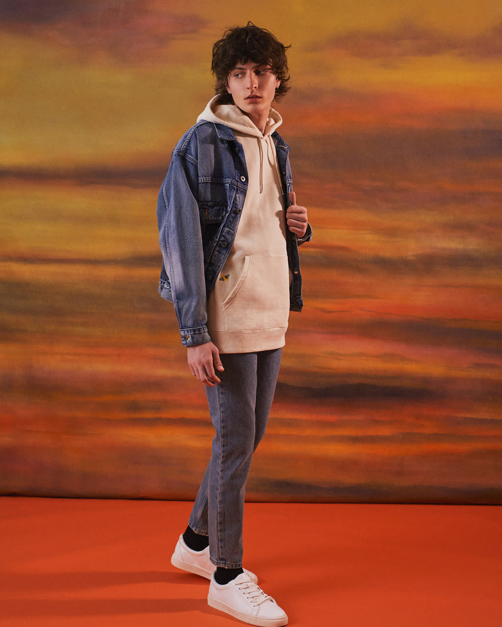 Person wearing a denim jacket over a Bee Embroidered Hoodie for Men with a sunset background