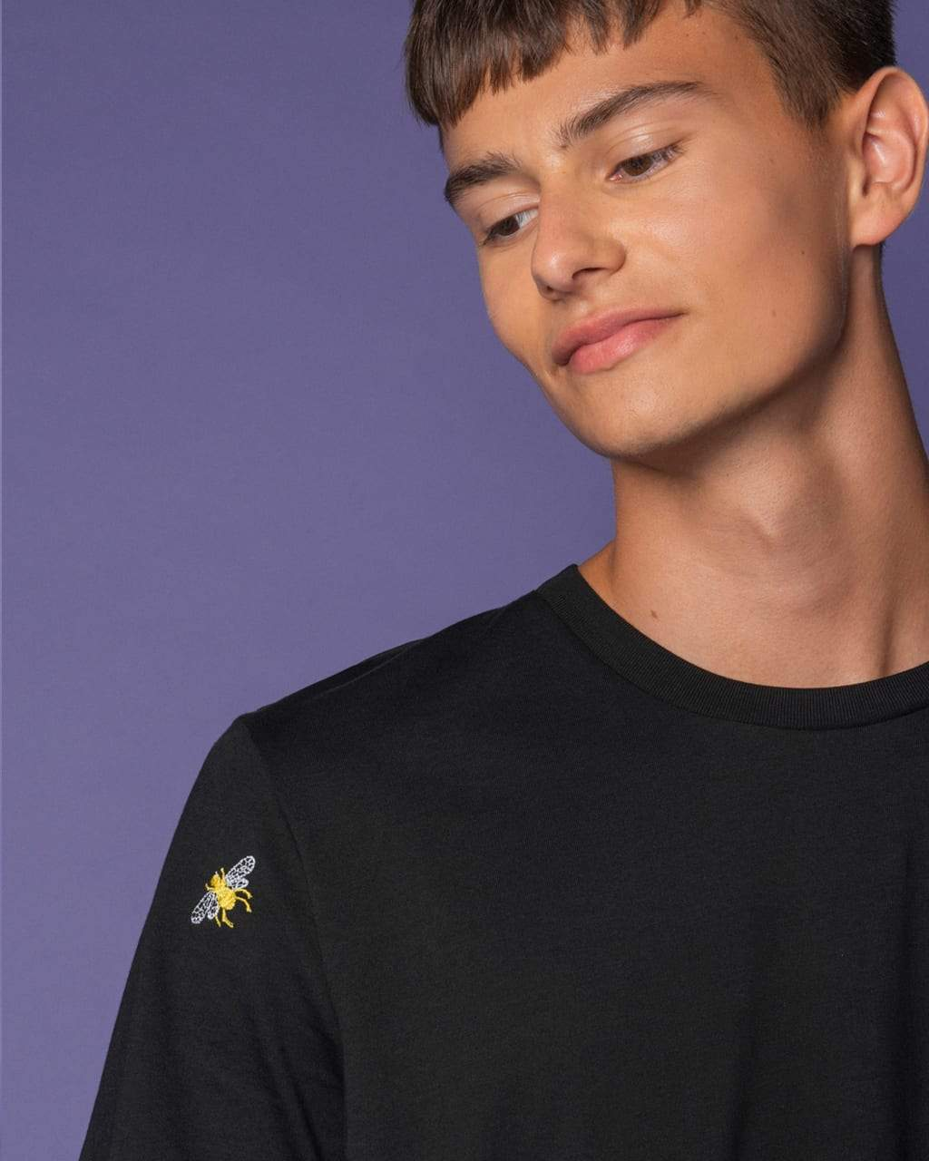 Person wearing a black Bee Embroidered Black T-Shirt with a small yellow graphic on a purple background