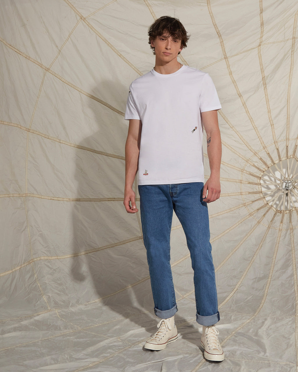 Man wearing a white t-shirt and blue jeans standing against a decorative wall.