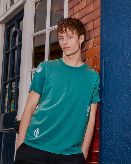 Man wearing a Shell Embroidered T-Shirt in Teal – 100% Organic Cotton, standing against a blue wall and brick wall.