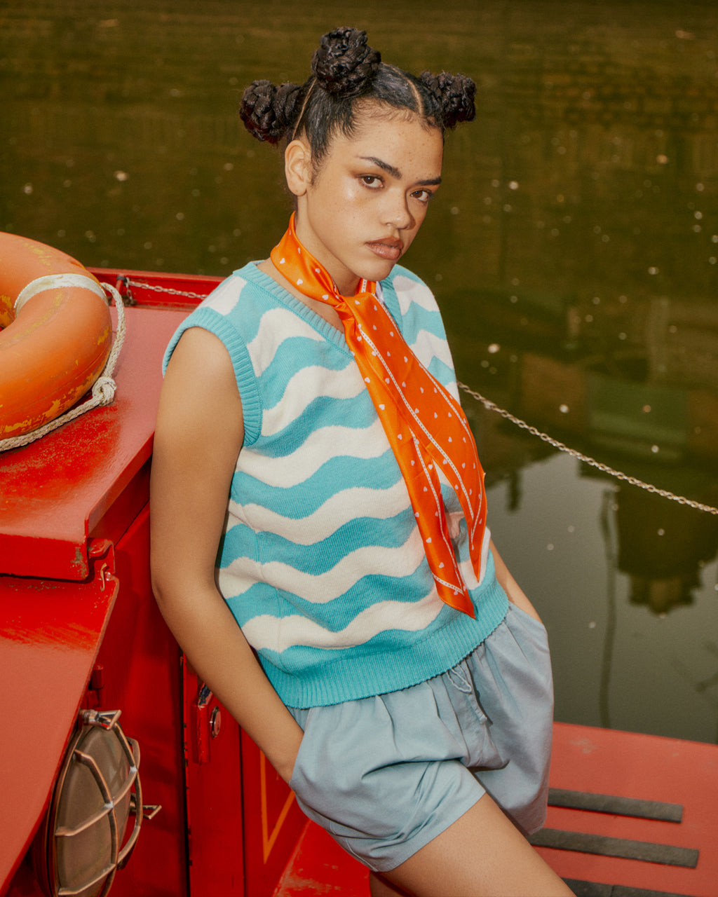 Woman wearing a Tiny Hearts Long Orange Silk Neck Scarf in a striped sweater vest and shorts standing on a red barge with water in the background